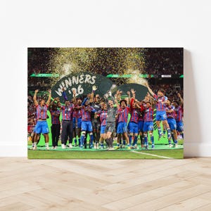 以下が含まれることがあります： サッカーチームが勝利を祝うキャンバスプリント。選手たちは青と赤のユニフォームを着てトロフィーを持ち、背後には「WINNERS '26」と書かれたバナーがあり、紙吹雪が舞っている。