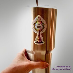 May include: A metallic, copper-toned insulated tumbler featuring a holographic sticker of a monstrance. The sticker displays a central IHS emblem, a cross, and radiating sunburst rays. A thin metal straw protrudes from the top of the tumbler.