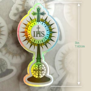 May include: A holographic sticker shaped like a monstrance featuring a central IHS emblem, a cross at the top, and a circular base with a Saint Benedict medal design. The sticker measures 7.62 cm in height and has a reflective, rainbow-coloured border.
