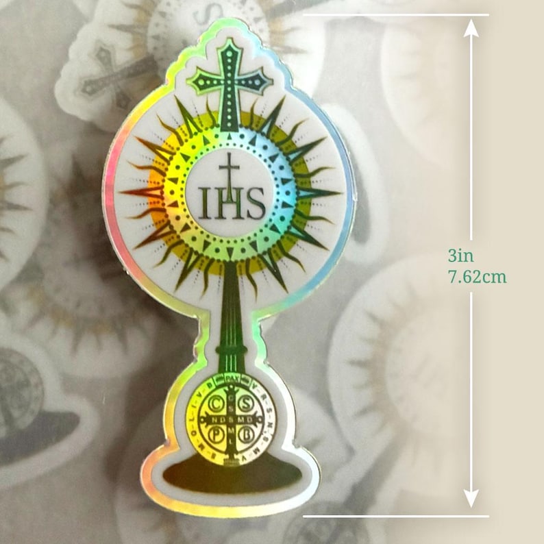May include: A holographic sticker shaped like a monstrance featuring a central IHS emblem, a cross at the top, and a circular base with a Saint Benedict medal design. The sticker measures 7.62 cm in height and has a reflective, rainbow-coloured border.