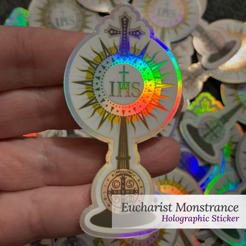 May include: A holographic sticker featuring a monstrance design with a central IHS emblem, a cross at the top, and a circular base containing a Saint Benedict medal motif. The sticker has a reflective, rainbow-coloured finish that catches light.