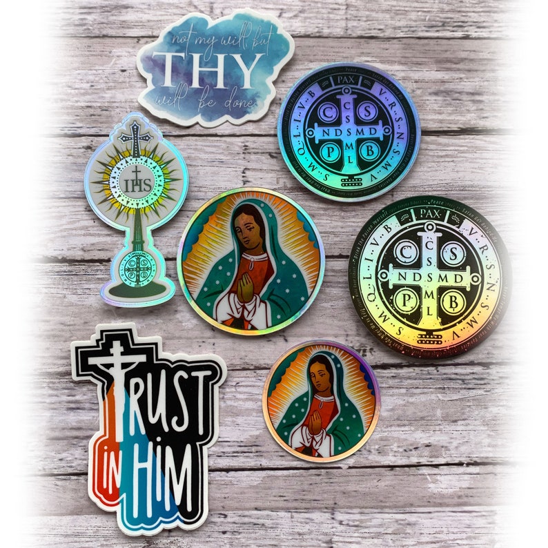 May include: A collection of seven religious stickers featuring two circular Saint Benedict medals with holographic finishes, two circular Our Lady of Guadalupe stickers, a monstrance sticker, and two die-cut stickers with the text Not my will but THY will be done and TRUST IN HIM.