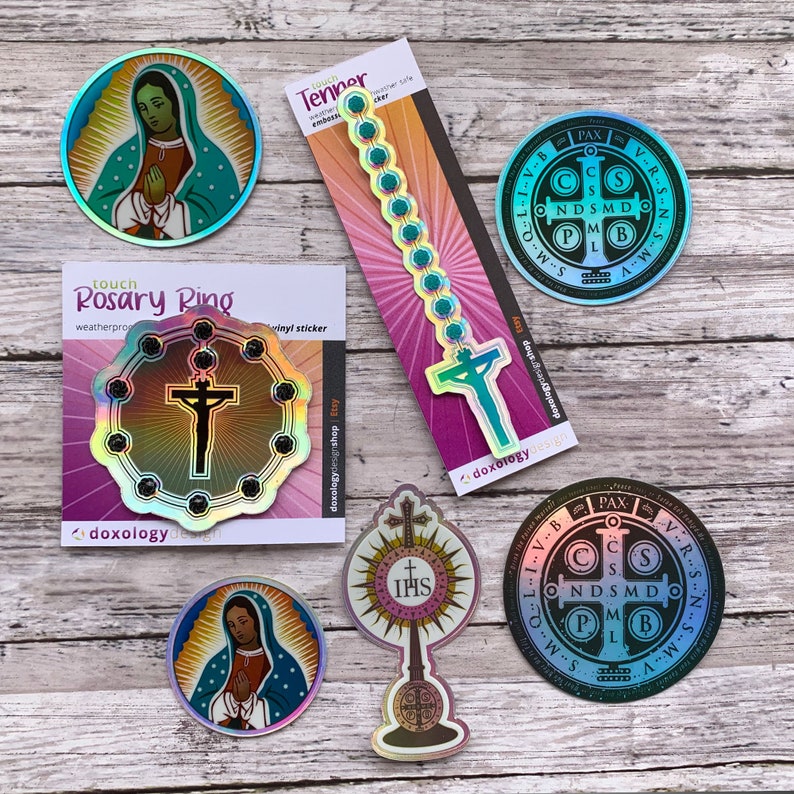 May include: A collection of religious stickers featuring holographic finishes. Included are two circular Our Lady of Guadalupe stickers, two circular Saint Benedict medals, a tenner rosary strip, a rosary ring sticker, and a monstrance sticker.