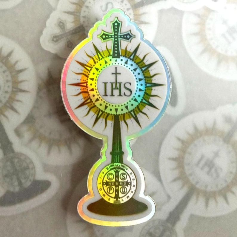 May include: A holographic sticker featuring a monstrance design with a central IHS emblem, a cross at the top, and a Saint Benedict medal at the base. The artwork uses green and white tones with a reflective rainbow border that catches the light.