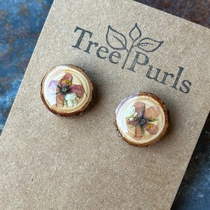 May include: A pair of round stud earrings featuring natural wood slices with pressed pink and white flowers encased in a clear resin finish. The earrings are mounted on a brown card printed with the text Tree Purls and Handmade in Oregon.