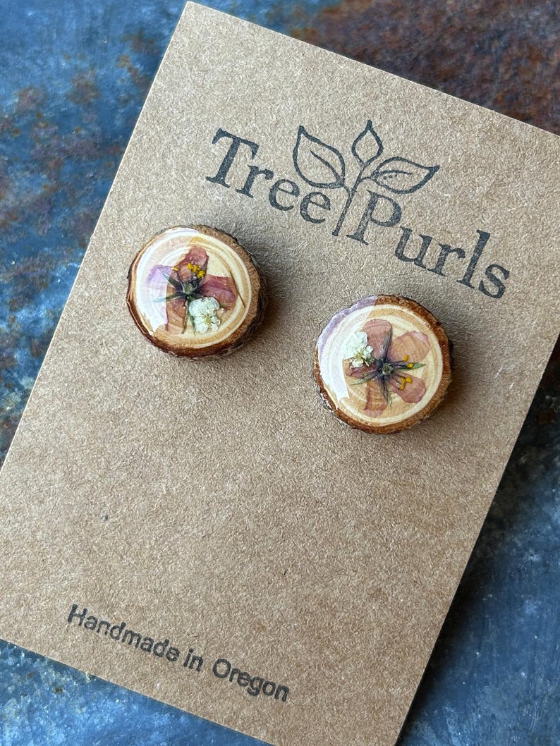 May include: A pair of round stud earrings featuring natural wood slices with pressed pink and white flowers encased in a clear resin finish. The earrings are mounted on a brown card printed with the text Tree Purls and Handmade in Oregon.