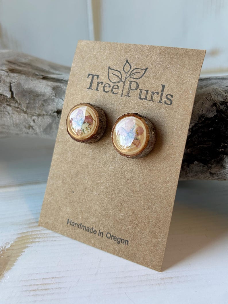 May include: A pair of round stud earrings made from natural wood slices with bark edges, featuring a glossy resin finish over a floral print. The earrings are attached to a brown card printed with the text Tree Purls and Handmade in Oregon.