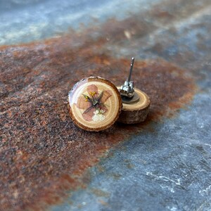 May include: A pair of stud earrings featuring circular slices of natural wood with bark edges. Each earring is topped with a clear resin coating over a pressed, dried flower with pink petals and yellow centers, attached to a metal post backing.