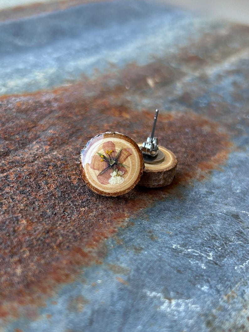 May include: A pair of stud earrings featuring circular slices of natural wood with bark edges. Each earring is topped with a clear resin coating over a pressed, dried flower with pink petals and yellow centers, attached to a metal post backing.