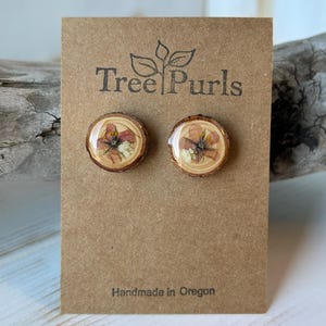 May include: A pair of round stud earrings featuring natural wood slices with pressed flowers encased in a clear resin finish. The earrings are mounted on a brown cardboard display card printed with the text Tree Purls and Handmade in Oregon.
