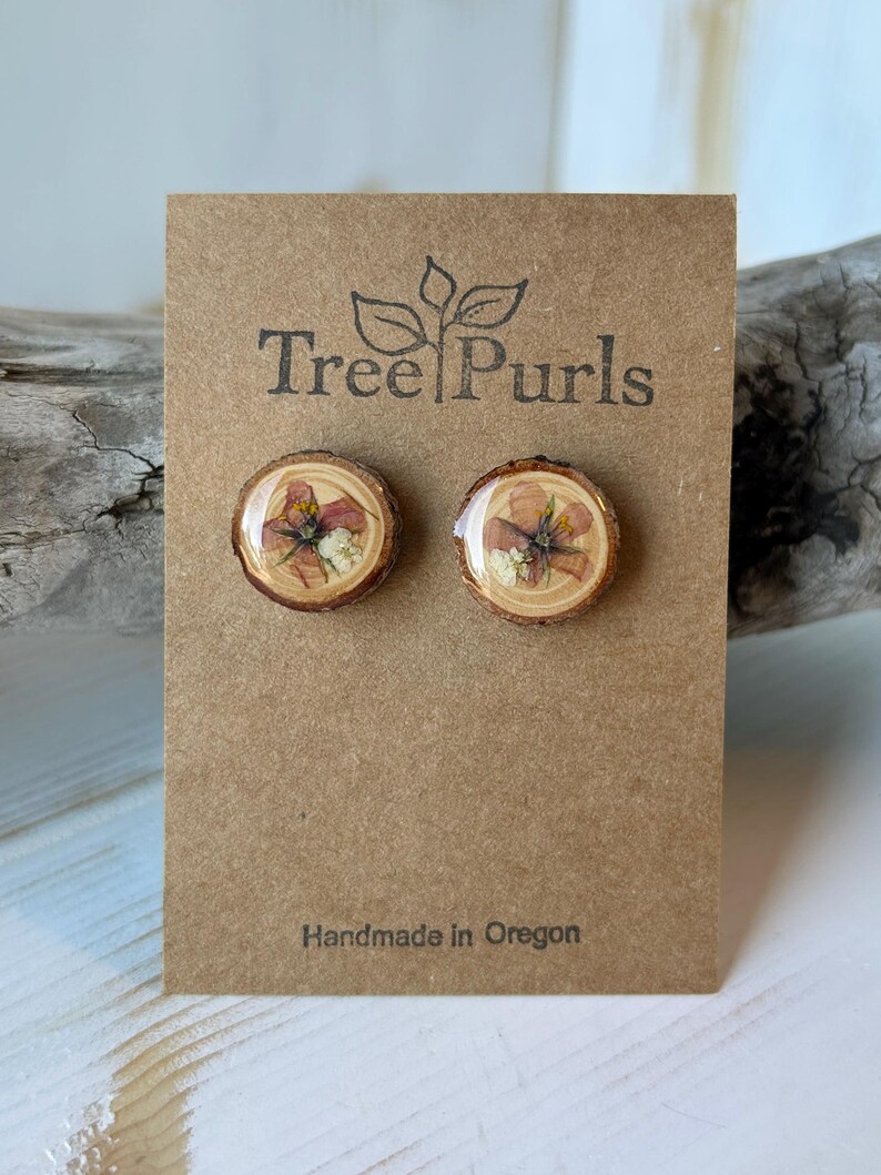 May include: A pair of round stud earrings featuring natural wood slices with pressed flowers encased in a clear resin finish. The earrings are mounted on a brown cardboard display card printed with the text Tree Purls and Handmade in Oregon.