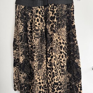 May include: A long skirt with a black leather-like waistband and a leopard print pattern in black and tan. The skirt is hanging on a gold hanger against a white background.