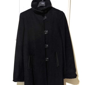 May include: A long, black coat with a high collar and three large, dark buttons down the front. The coat has two pockets and a subtle sheen, suggesting a wool or wool-blend fabric. The coat is hanging on a white door.