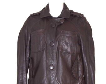 Könnte beinhalten: Dunkelbraune Lederjacke mit klassischem Design. Die Jacke hat einen Kragen, eine Knopfleiste, zwei Brusttaschen und geknöpfte Manschetten. Die Jacke wird auf einer weißen Schaufensterpuppe präsentiert.