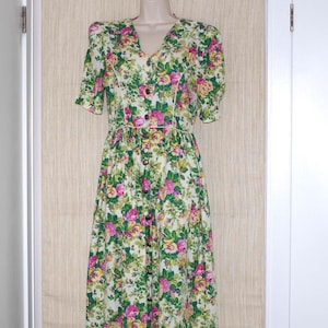 Puede incluir: Vestido con estampado floral, escote en V y mangas cortas. El vestido tiene una botonadura frontal y una falda amplia. El estampado incluye flores rosas, amarillas y verdes sobre fondo blanco. El vestido se exhibe en un maniquí.