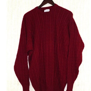 May include: A burgundy cable-knit sweater with long sleeves, hanging on a wooden hanger. The sweater has a crew neck and a textured pattern. The label on the sweater is white with black text.