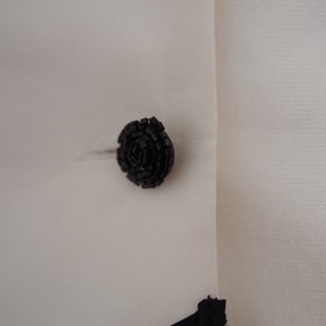 May include: Close-up of a black, circular button on a white garment. The button is made of small, textured beads arranged in a spiral pattern. The fabric has a subtle texture.