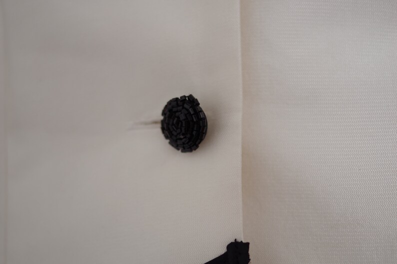 May include: Close-up of a black, circular button on a white garment. The button is made of small, textured beads arranged in a spiral pattern. The fabric has a subtle texture.