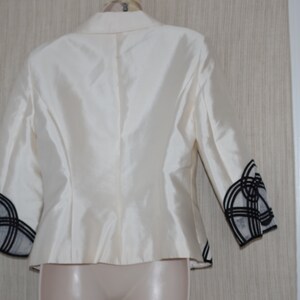May include: Ivory-colored blazer with three-quarter sleeves. The sleeves feature a black and white geometric design at the cuffs. The jacket has a classic tailored fit and is made of a shiny, possibly silk, fabric.