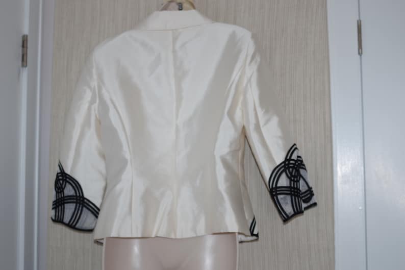 May include: Ivory-colored blazer with three-quarter sleeves. The sleeves feature a black and white geometric design at the cuffs. The jacket has a classic tailored fit and is made of a shiny, possibly silk, fabric.
