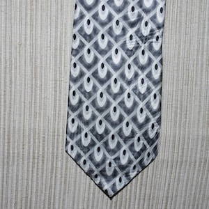 May include: A patterned necktie featuring a repeating design of white, gray, and black oval shapes within a diamond grid. The tie is angled, showcasing its geometric pattern against a neutral background.