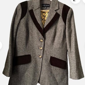 May include: A grey tweed blazer with dark brown velvet detailing on the collar, shoulders, and pockets. The jacket features three gold buttons and a patterned lining. The label reads "SANDY KEMPER".