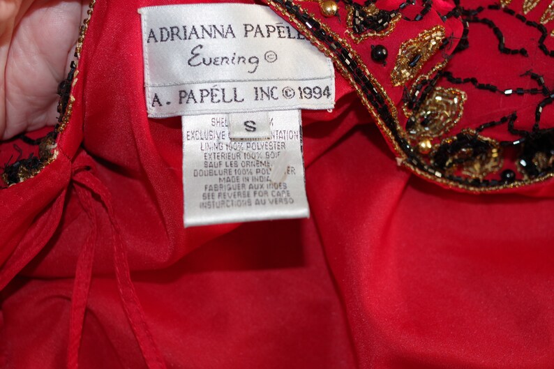May include: Red evening dress with black and gold beaded embellishments. The label reads "ADRIANNA PAPELL Evening" and "A. PAPELL INC &copy; 1994". The dress is size S and made in India.