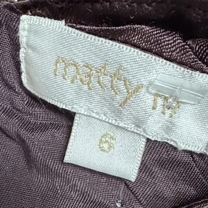 May include: Close-up of a clothing label with the brand name "matty" in gold lettering. The label is white with a size 6 tag below it. The fabric is a dark purple colour.