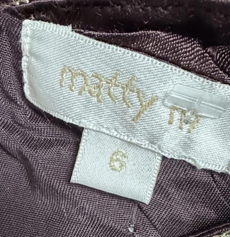 May include: Close-up of a clothing label with the brand name "matty" in gold lettering. The label is white with a size 6 tag below it. The fabric is a dark purple colour.