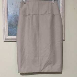 May include: A light beige pencil skirt hangs on a hanger. The skirt is knee-length and has a high waist with a structured design. The fabric appears smooth and has a slight sheen. The skirt is a neutral color, suitable for various outfits.