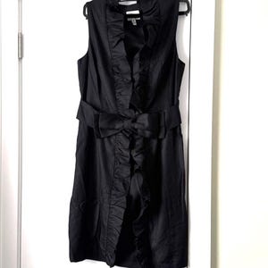 May include: A sleeveless black dress with a ruffled front and a bow belt. The dress hangs on a black hanger against a white door. The dress has a knee-length hemline and a satin-like sheen.