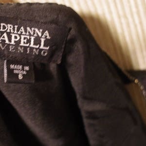 May include: A close-up shot of a black garment tag displaying the brand name "ADRIANNA PAPELL EVENING" in white. Beneath, a smaller tag reads "MADE IN INDIA" and the size "8". The garment is likely a dress or evening wear, possibly a formal gown.