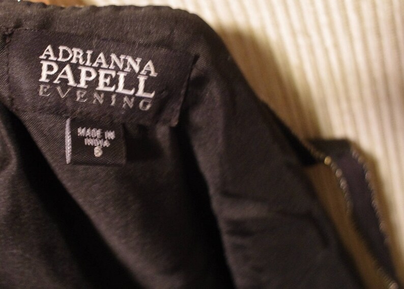 May include: A close-up shot of a black garment tag displaying the brand name "ADRIANNA PAPELL EVENING" in white. Beneath, a smaller tag reads "MADE IN INDIA" and the size "8". The garment is likely a dress or evening wear, possibly a formal gown.