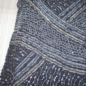 May include: Detailed view of a dark blue garment adorned with beads and sequins. The design incorporates diagonal stripes and wavy lines in silver and grey, producing a textured, shimmering appearance. The fabric seems to be draped.
