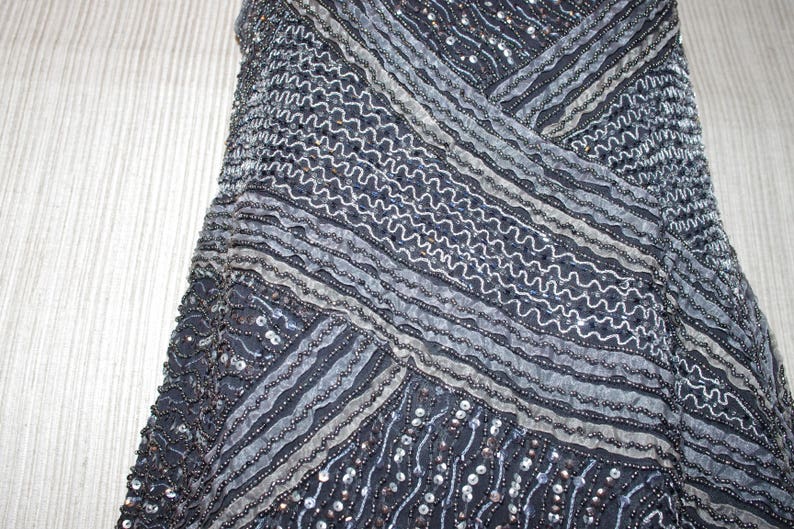 May include: Detailed view of a dark blue garment adorned with beads and sequins. The design incorporates diagonal stripes and wavy lines in silver and grey, producing a textured, shimmering appearance. The fabric seems to be draped.