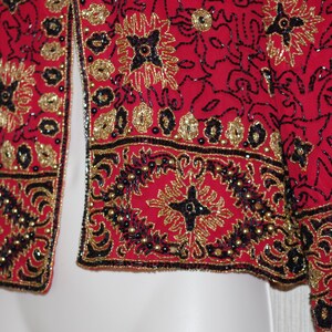 May include: A red jacket with intricate gold and black beadwork. The jacket features floral and geometric patterns, with a beaded border along the edges. The design includes gold sequins and black beads, creating a detailed and ornate look.