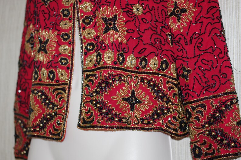 May include: A red jacket with intricate gold and black beadwork. The jacket features floral and geometric patterns, with a beaded border along the edges. The design includes gold sequins and black beads, creating a detailed and ornate look.