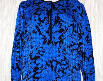 Puede incluir: Una blusa de manga larga con un vibrante estampado floral azul sobre un fondo negro. La blusa tiene un cuello redondo, una botonadura frontal y ribetes negros en el cuello, los puños y el dobladillo. La tela parece tener una textura sedosa.
