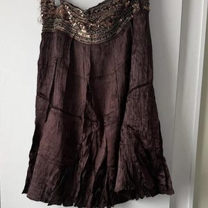 May include: A dark brown skirt with a tiered design and a decorative waistband. The waistband is embellished with sequins and intricate embroidery in gold and brown. The skirt has a flowing, textured appearance.