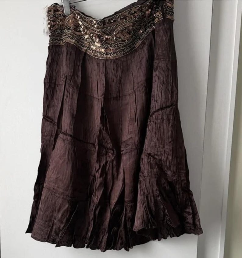 May include: A dark brown skirt with a tiered design and a decorative waistband. The waistband is embellished with sequins and intricate embroidery in gold and brown. The skirt has a flowing, textured appearance.