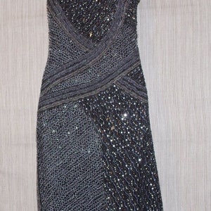 May include: A dark grey, beaded evening dress with spaghetti straps. The dress has a V-neckline and a fitted silhouette. The bodice and skirt are embellished with sequins and beads, creating a shimmering effect. The dress is displayed on a mannequin.