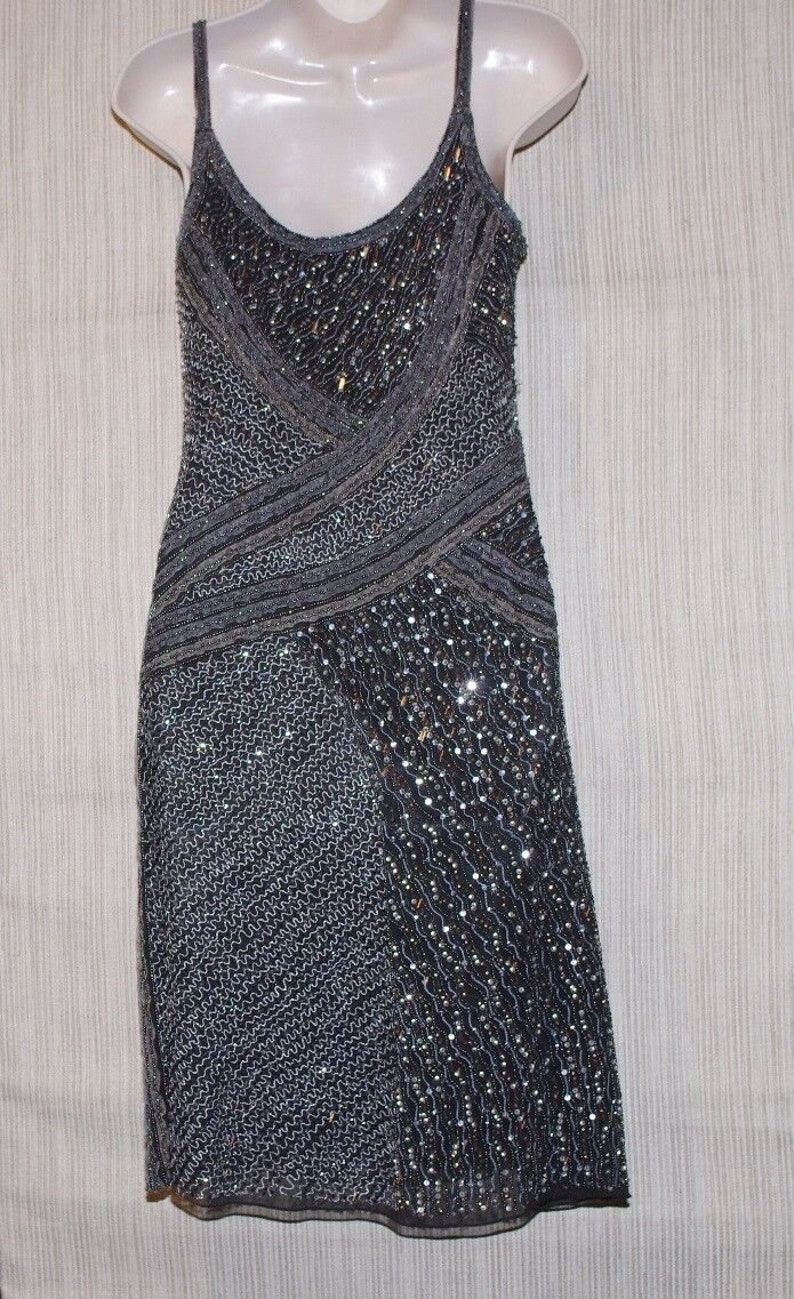 May include: A dark grey, beaded evening dress with spaghetti straps. The dress has a V-neckline and a fitted silhouette. The bodice and skirt are embellished with sequins and beads, creating a shimmering effect. The dress is displayed on a mannequin.
