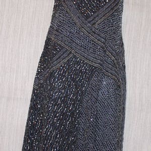 May include: A dark grey, sleeveless dress with a beaded and sequined design. The dress features a crisscross pattern across the bodice and a flowing skirt. The dress is displayed on a white mannequin.