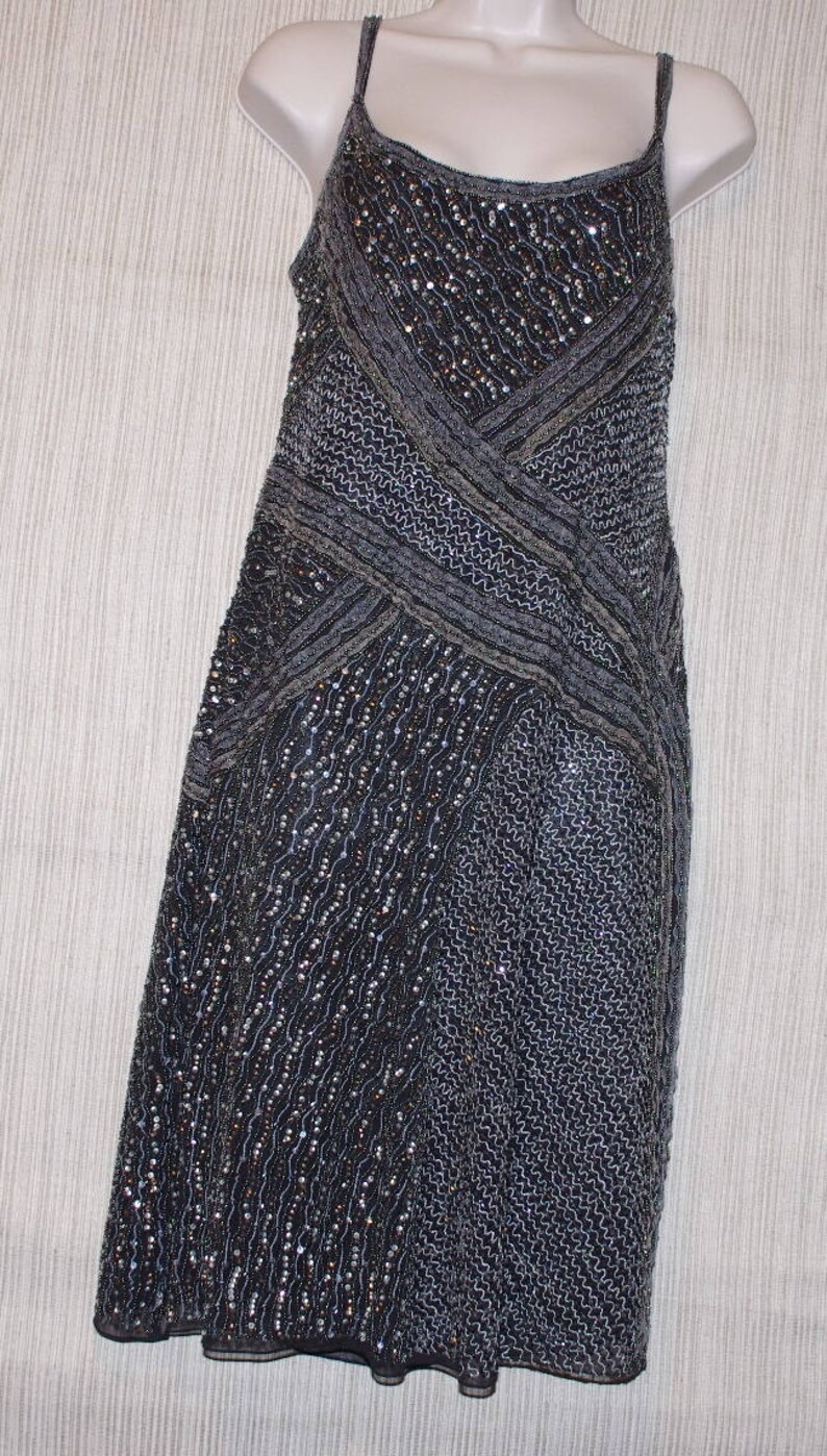 May include: A dark grey, sleeveless dress with a beaded and sequined design. The dress features a crisscross pattern across the bodice and a flowing skirt. The dress is displayed on a white mannequin.