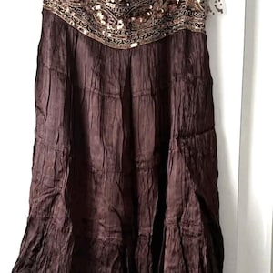 May include: A brown tiered skirt, reaching the knee. The waistband is adorned with gold sequins and beadwork. The fabric has a crinkled texture. The skirt is suspended from a hanger. The skirt's length is approximately 60 cm.