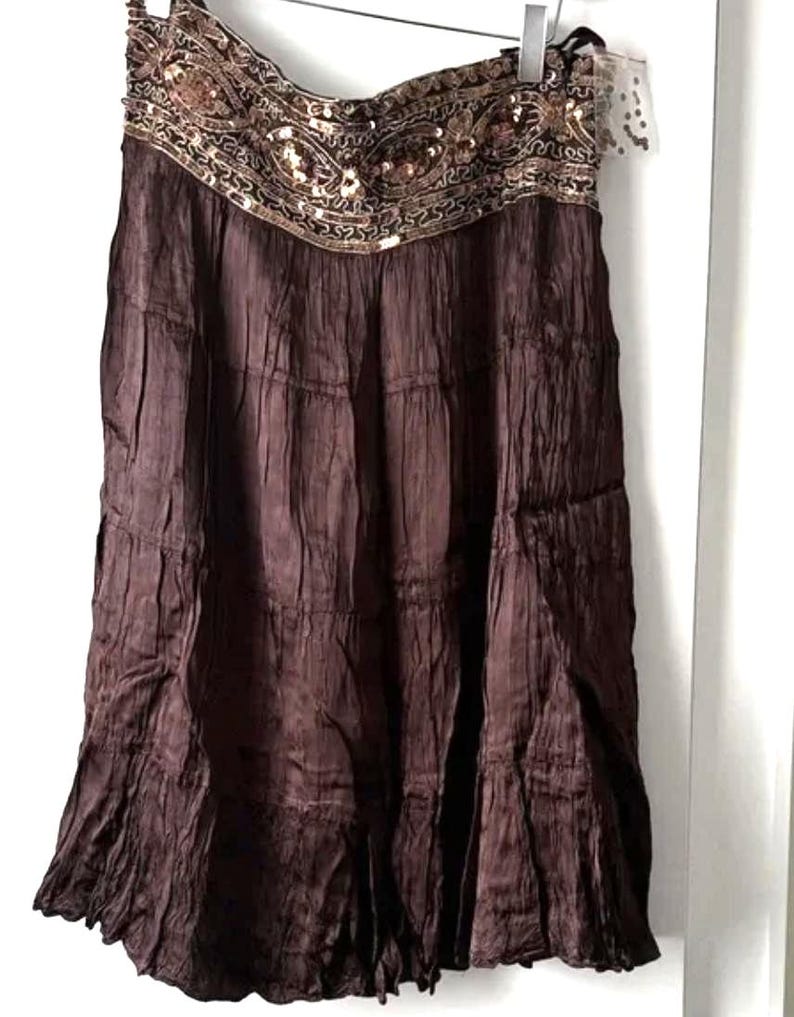 May include: A brown tiered skirt, reaching the knee. The waistband is adorned with gold sequins and beadwork. The fabric has a crinkled texture. The skirt is suspended from a hanger. The skirt's length is approximately 60 cm.