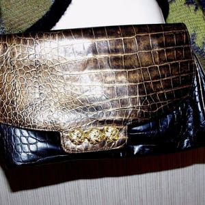 Peut inclure: Pochette bicolore avec un motif imitation peau de crocodile. Le rabat supérieur est marron et doré, tandis que le bas est noir. La pochette a une garniture dorée et une forme rectangulaire.