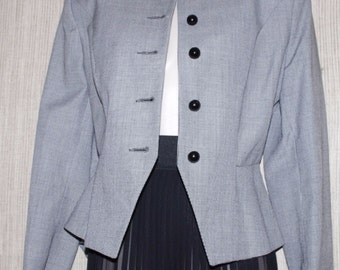 May include: A light grey blazer with a small checked pattern, featuring a round collar and black buttons. The blazer is paired with a dark skirt. The jacket has three buttons on the left side and four on the right.