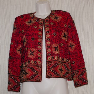 May include: A red jacket with intricate gold and black beadwork. The jacket features a round neckline and long sleeves. The design includes floral and geometric patterns, creating a decorative and elegant look. The jacket is displayed on a mannequin.