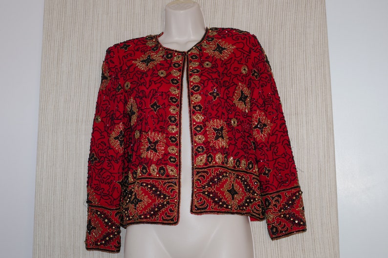 May include: A red jacket with intricate gold and black beadwork. The jacket features a round neckline and long sleeves. The design includes floral and geometric patterns, creating a decorative and elegant look. The jacket is displayed on a mannequin.